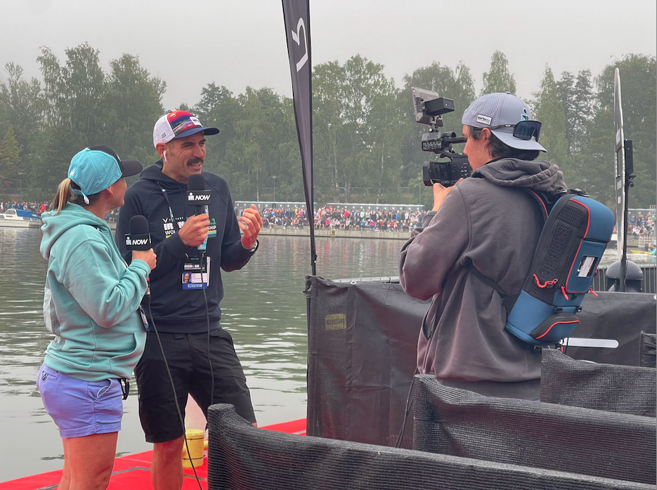 Camera Operator takes video of 2 Sports Commentators in front of a lake.